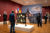 A large mural of a church and cemetery is in the middle of an art gallery, filled with people looking at the art on walls and artwork standing independently.